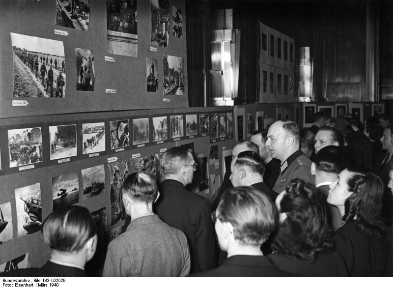 Ausstellung des NS-Reichsausschusses der Bildberichterstatter, Berlin März 1940, Fotograf: Eisenhart Original-Bildunterschrift ADN: „Bildberichterstatter stellen aus. Der faschistische Reichsausschuss der Bildberichterstatter im Reichsverband der Deutschen Presse veranstaltete Mitte März 1940 im Rahmen eines Kulturabends eine Ausstellung von Spitzenleistungen seiner Mitglieder unter besonderer Brücksichtigung der Arbeit der Angehörigen der Propaganda-Kompanie der faschistischen deutschen Wehrmacht.- Blick in die Ausstellung. (Mitte in Wehrmachtsuniform) der Leiter des Referats "Bildpresse" im faschistischen Propagandaminsteriums, Heiner Kurzbein.“ Quelle: Bundesarchiv Wikimedia Comons, Lizenz: CC