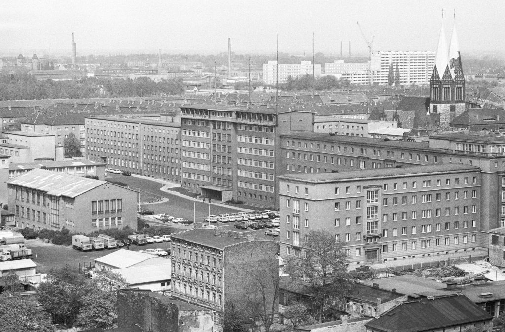 „Wo Mielke Dienst tut. Berlin (Bezirk Berlin) DDR, 26. 04. 1974. Karree des Staatssicherheitsministeriums (MfS). Die Dienstgebaeude des Ministeriums fuer Staatssicherheit (MfS, Stasi) befinden sich an der Ecke Normannenstrasse, Frankfurter Allee. Der Minister residiert in den oberen Stockwerken des erhabenen Teils des Hauptgebaeudes. Von der SED wird der Geheimdienst als Schwert und Schild der Partei bezeichnet. Das Volk belegt die Behoerde mit Begriffen wie Firma; Guck, Horch und Greif.“ Foto Klaus Mehner © Bundesstiftung Aufarbeitung, Klaus Mehner, 74_0426_POL_MfS-DG_02 mit freundlicher Genehmigung