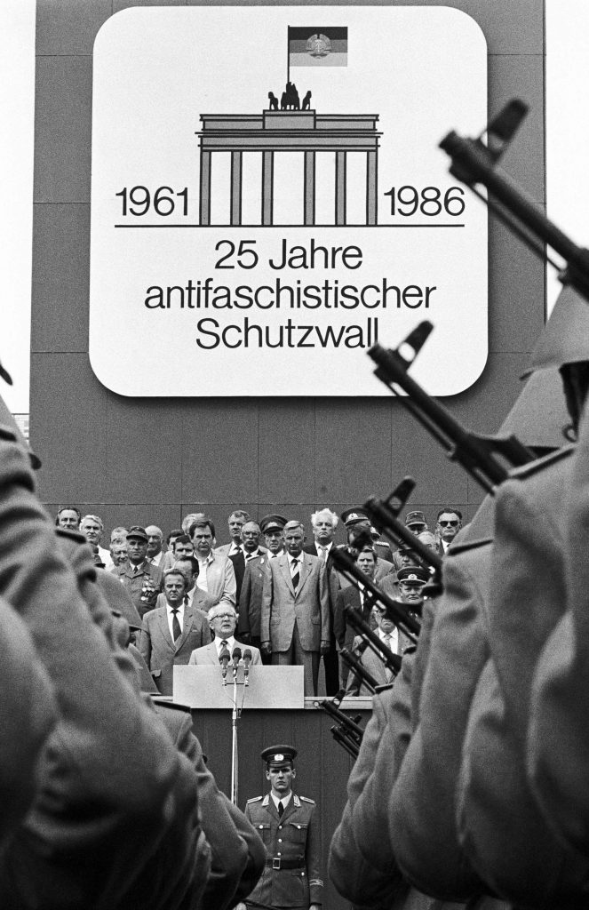 „Berlin (Bezirk Berlin) DDR, 13-08-1986. Mit einem Kampfappell der Kampfgruppen (KG) der Arbeiterklasse beging die Deutsche Demokratische Republik (DDR) den 25. Jahrestag des Baus des Antifaschistischen Schutzwalls, vulgo Mauer. Der SED-Chef würdigte das Bauwerk als Voraussetzung für die Friedens- und Entspannungspolitik. Alsdann nahmen Partei- und Staatsführung den Vorbeimarsch von Soldaten der Nationalen Volksarmee (NVA) und Betriebskampfgruppen ab. Erich Honecker (SED) bei Festrede.“ Foto: Klaus Mehner © Bundesstiftung Aufarbeitung, Klaus Mehner, 86_0813_POL_25JT_Mauer_17