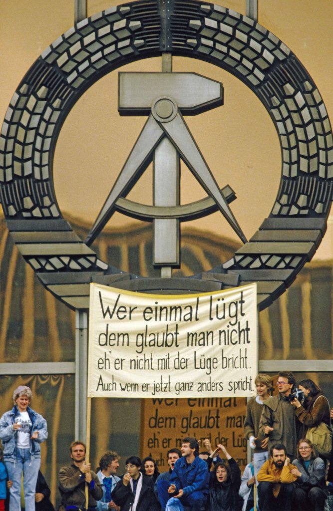 „Massendemo läutet Ende der SED-Diktatur ein. Berlin (Bezirk Berlin) DDR, 04.11.1989. Demonstranten mit Transparent, DDR-Signet am Palast der Republik. Mit ihrem Alleinvertretungsanspruch ist die SED am Ende, die Existenz des gesamten Regimes und der DDR steht auf der Kippe. Mehr als eine Million Menschen demonstrieren für Presse-, Meinungs- und Versammlungsfreiheit im Stadtzentrum und auf dem Alexanderplatz.“ Foto: Klaus Mehner© Bundesstiftung Aufarbeitung, Klaus Mehner, 89_1104_POL-Demo_40